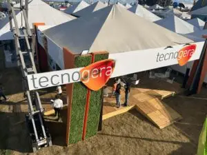 No estande da Tecnogera na Bahia Farm Show, grandes tendas brancas e uma placa verde e laranja com o nome da empresa chamam a atenção. Os visitantes circularam pelo deque de madeira sobre a grama seca, conhecendo as oportunidades do agronegócio. Mesas e cadeiras ficam dispostas sob as tendas, enquanto o sol forte ilumina o espaço e projeta sombras nítidas ao redor.