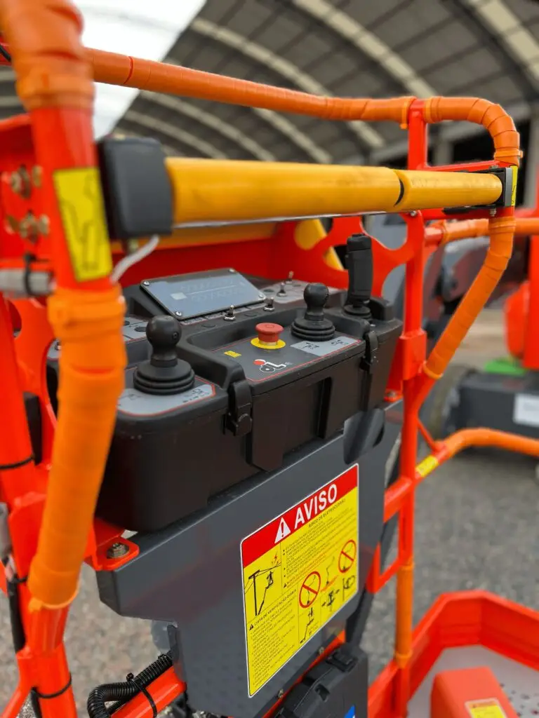 Close da mesa de controle de uma tesoura elevatória laranja da Tecnogera, com dois joysticks, vários botões e sensor antiesmagamento para maior segurança. Em destaque, um aviso amarelo com a palavra "AVISO" em vermelho, tudo sobre um fundo industrial desfocado.