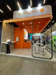 O estande da Tecnogera na Brazil Windpower chama a atenção com um design moderno em laranja e branco, equipado com balcão, banquetas e telas digitais. Uma mulher aparece próxima ao balcão segurando uma bolsa, enquanto outras pessoas passam pelo local, evidenciando o foco em energias renováveis. O ambiente é iluminado por luzes brilhantes no teto.