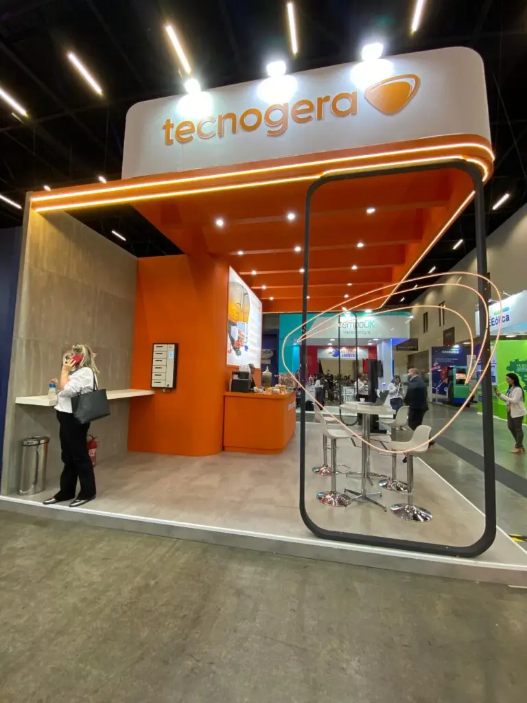 O estande da Tecnogera na Brazil Windpower chama a atenção com um design moderno em laranja e branco, equipado com balcão, banquetas e telas digitais. Uma mulher aparece próxima ao balcão segurando uma bolsa, enquanto outras pessoas passam pelo local, evidenciando o foco em energias renováveis. O ambiente é iluminado por luzes brilhantes no teto.