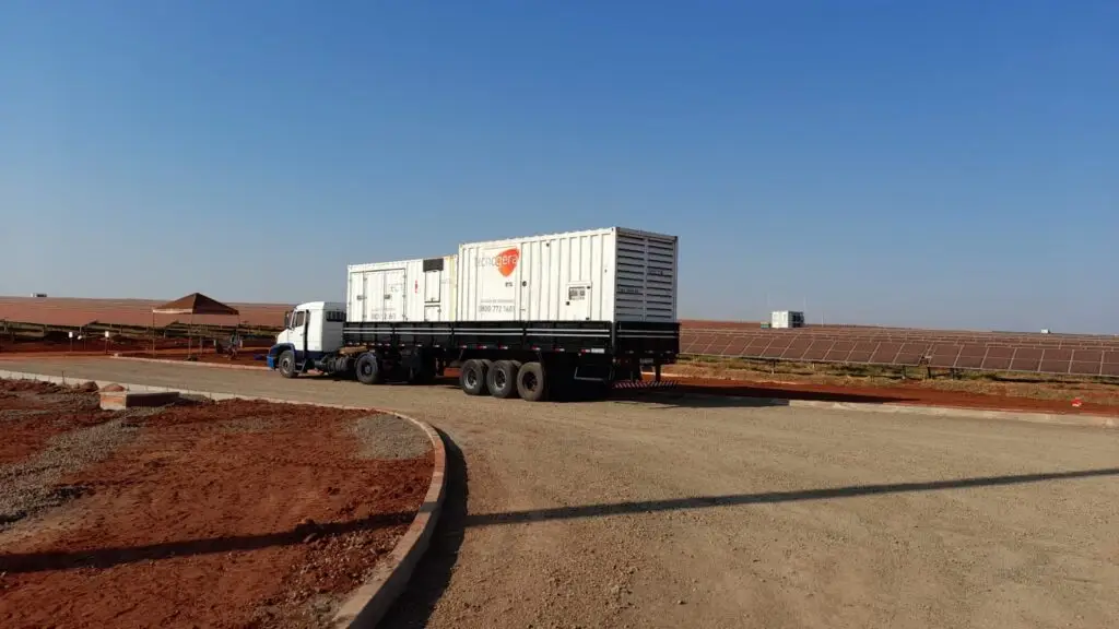 Um caminhão branco com carreta prancha transporta dois grandes contêineres brancos e está estacionado em uma estrada de terra e cascalho. Ao fundo, há fileiras de painéis solares instaladas sobre o solo avermelhado, reforçando o compromisso da Tecnogera com soluções de energia para a gestão do agronegócio sob um céu azul claro.
