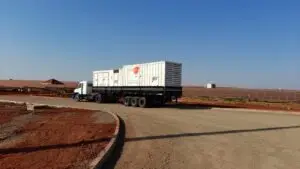 Um caminhão branco com carreta prancha transporta dois grandes contêineres brancos e está estacionado em uma estrada de terra e cascalho. Ao fundo, há fileiras de painéis solares instaladas sobre o solo avermelhado, reforçando o compromisso da Tecnogera com soluções de energia para a gestão do agronegócio sob um céu azul claro.