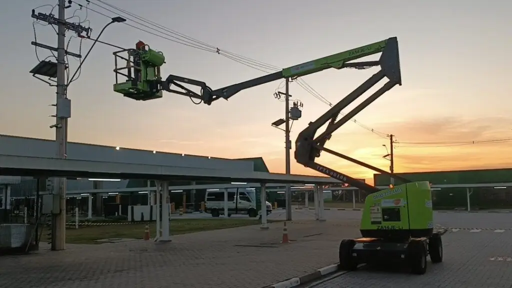 Um colaborador, usando colete refletivo, opera uma plataforma elevatória articulada verde da Tecnogera para acessar cabos de energia em um poste ao entardecer. O veículo está estacionado em um pátio pavimentado próximo a uma estrutura coberta, sinalizado por cones laranja.