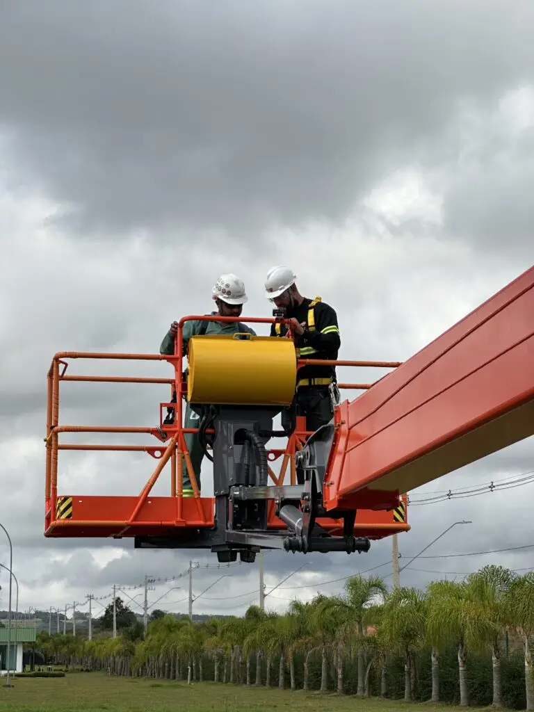 Dois profissionais da Tecnogera, usando capacetes e cintos de segurança, estão sobre uma plataforma elevatória laranja. Eles demonstram habilidade no manuseio do equipamento enquanto inspecionam ou conversam sobre alguma atividade no canteiro de obras ao ar livre, sob um céu nublado.