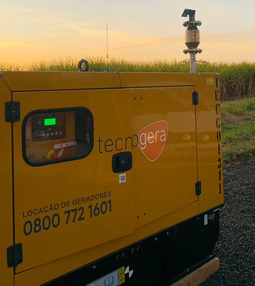 Um gerador de energia amarelo da Tecnogera está posicionado ao ar livre, sobre um piso de cascalho, próximo a uma área de vegetação alta, durante o pôr do sol. Exibindo painel digital, tubo de ventilação e o telefone 0800 772 1601 em destaque, ele representa a eficiência operacional da Tecnogera sob um céu com tons alaranjados e azulados.