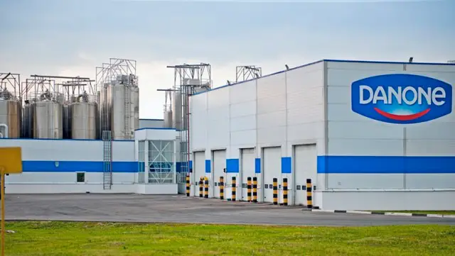 A imagem mostra uma moderna fábrica da Danone Alimentos, com paredes brancas e detalhes em azul. Grandes silos metálicos aparecem ao fundo, destacando o logotipo da Danone. Na frente do prédio, há uma área pavimentada com um pouco de grama e o céu está nublado. Esta é mais uma solução de energia temporária atendida pela Tecnogera.
