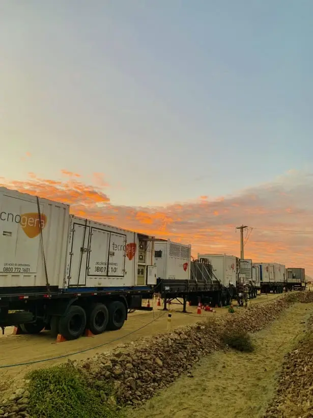 Vários caminhões brancos, identificados com a marca Tecnogera, empresa especialista em geradores a diesel, estão estacionados lado a lado em uma estrada de terra ao entardecer. O céu alaranjado realça as nuvens sobre o terreno rochoso e seco que cerca a via.