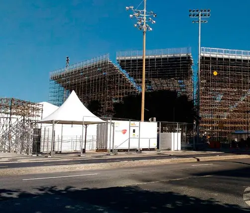 Uma grande arquibancada metálica, parte da construção para as Olimpíadas Rio 2016, destaca-se sob um céu azul e sem nuvens. Na frente dela, vemos tendas brancas, um prédio móvel, cercas e uma estrada com faixas amarelas. Altos postes de luz completam o cenário ao redor da estrutura. Esta é mais uma operação que conta com a expertise da Tecnogera.