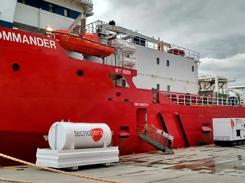 Um grande navio vermelho chamado "COMMANDER" está atracado no porto em um dia nublado. No píer, um tanque cilíndrico branco com o logotipo da Tecnogera garante uma solução de energia eficiente e econômica, estando seguro por cabos perto da embarcação de vários andares.