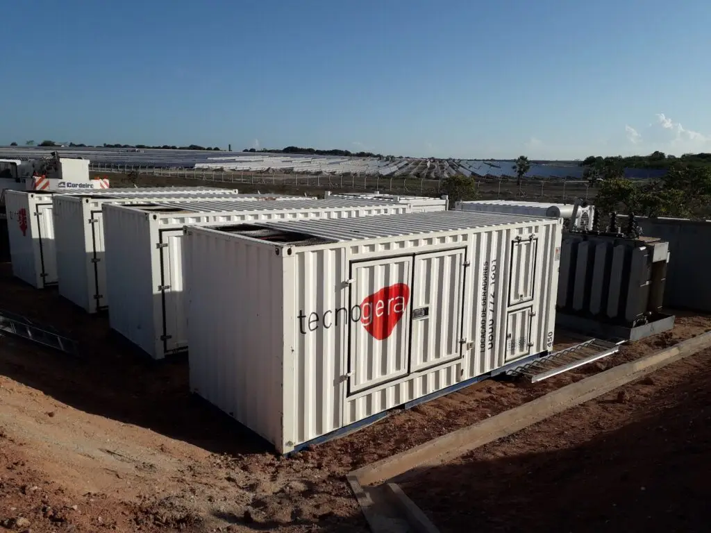 Diversos geradores de energia brancos da Tecnogera, acomodados em contêineres, estão posicionados sobre um solo avermelhado próximo a uma usina solar. Ao fundo, vêem-se fileiras de painéis solares e áreas verdes sob um céu azul, mostrando que a Tecnogera está pronta para fornecer energia em Stand By sempre que necessário.