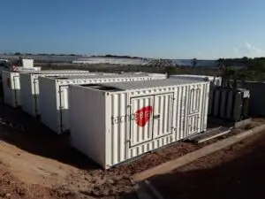 Diversos geradores de energia brancos da Tecnogera, acomodados em contêineres, estão posicionados sobre um solo avermelhado próximo a uma usina solar. Ao fundo, vêem-se fileiras de painéis solares e áreas verdes sob um céu azul, mostrando que a Tecnogera está pronta para fornecer energia em Stand By sempre que necessário.