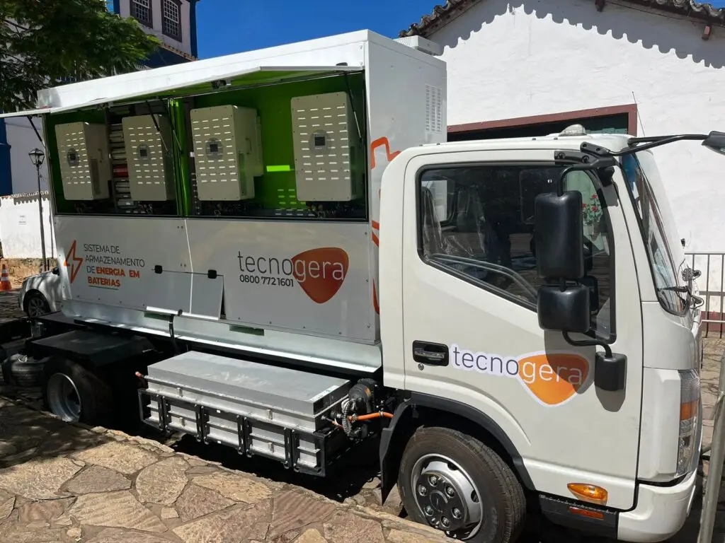 Um caminhão branco da Tecnogera está estacionado em uma rua de paralelepípedos, com a lateral aberta mostrando painéis de controle elétrico em seu interior. Exibindo o logotipo e as informações de contato da Tecnogera, a imagem destaca oportunidades no mercado de acesso, tendo ao fundo prédios banhados pelo sol.