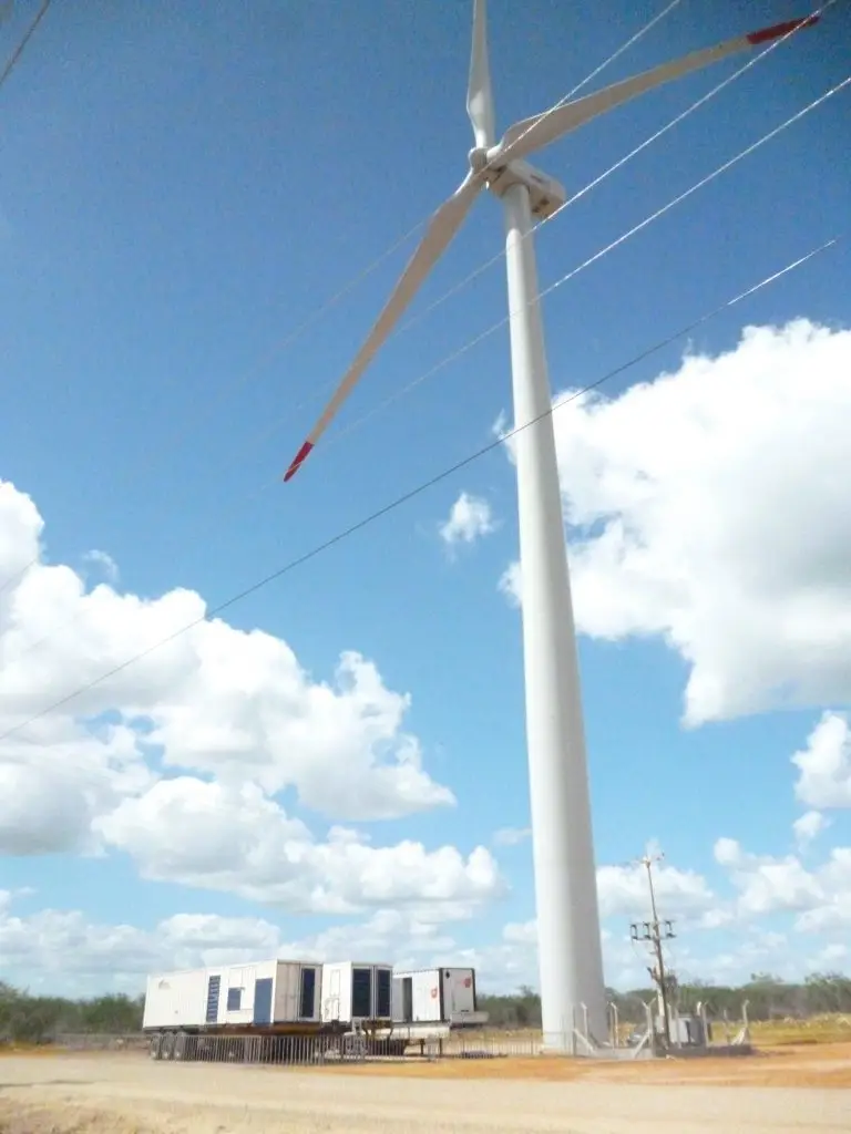 Uma turbina eólica branca e alta, com três pás, se destaca em meio ao céu azul com algumas nuvens, representando a energia limpa do vento. No solo seco abaixo, equipamentos elétricos e transformadores estão posicionados próximos, enquanto linhas de transmissão cortam a imagem na horizontal. Essa é mais uma demonstração das soluções fornecidas pela Tecnogera.