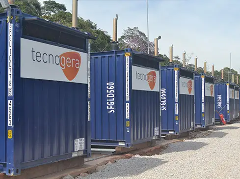 Diversos contêineres azuis com a marca "Tecnogera" estão alinhados sobre um caminho de cascalho, compondo parte de uma usina termelétrica. Cada contêiner possui um número de identificação e hastes verticais em seu topo. Ao fundo, é possível ver árvores e o céu nublado.