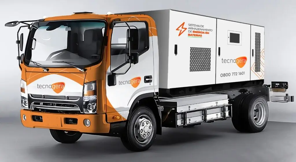 Um caminhão de médio porte da Tecnogera, com cabine laranja e branca, transporta uma unidade compacta de armazenamento de energia em sua carroceria. O equipamento conta com detalhes como entradas de ar, uma porta e avisos sobre a reutilização de baterias. O veículo está estacionado sobre um fundo claro e neutro.