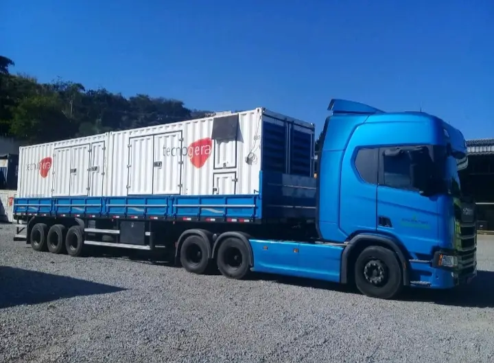 Na imagem, um caminhão azul com uma carreta plataforma transporta dois grandes contêineres brancos UGTEM da Tecnogera, identificados pelo nome da empresa e por logotipos de corações vermelhos. O veículo está estacionado em um terreno de cascalho, sob um céu azul e claro, com árvores e algumas construções ao fundo.