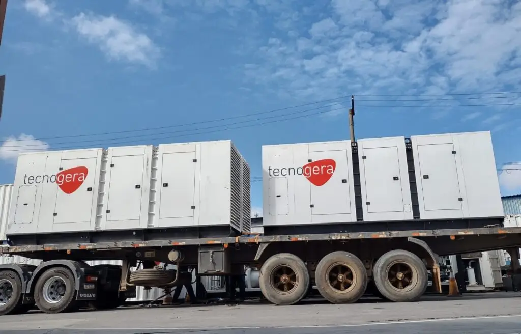 Um caminhão plataforma transporta dois grandes geradores de energia brancos, identificados com o nome Tecnogera e um logotipo de coração vermelho. O veículo está estacionado ao ar livre, sob um céu azul com poucas nuvens, ilustrando as vantagens do aluguel de geradores. Ao fundo, é possível ver linhas de energia elétrica.