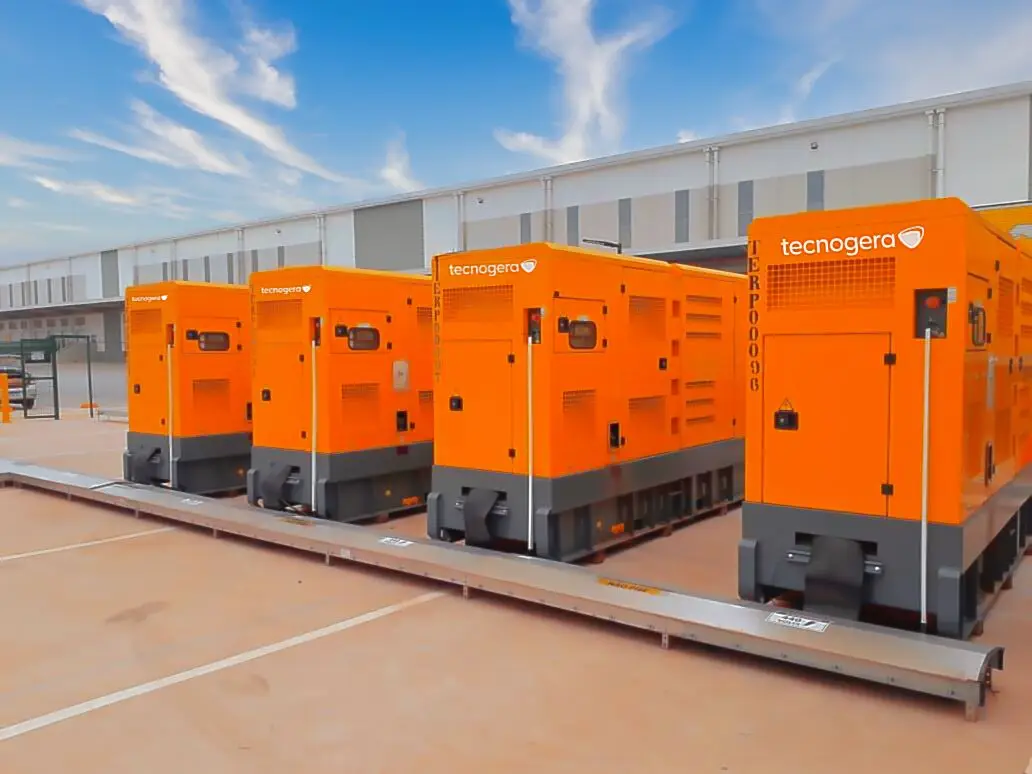 Quatro grandes geradores industriais laranja, com o nome Tecnogera, estão alinhados ao ar livre sobre plataformas metálicas para trabalho em altura, próximos a um moderno galpão cinza-claro. O céu parcialmente azul com algumas nuvens completa o cenário.