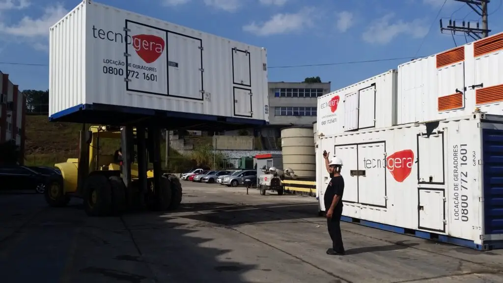 Uma empilhadeira amarela ergue um grande contêiner branco com a marca Tecnogera, enquanto um trabalhador com capacete orienta a operação ao lado. Ao fundo, aparecem outros contêineres, carros estacionados e plataformas para trabalhos em altura, tudo sob um céu azul.