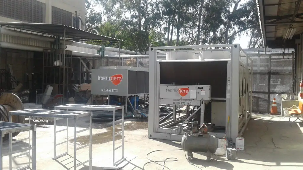 Área industrial ao ar livre com grandes geradores prateados identificados com a marca Tecnogera. O espaço conta também com estruturas metálicas, cabos, um compressor de ar, cones de trânsito laranja e branco, plataformas para trabalho em altura e árvores altas ao fundo sob luz solar intensa.