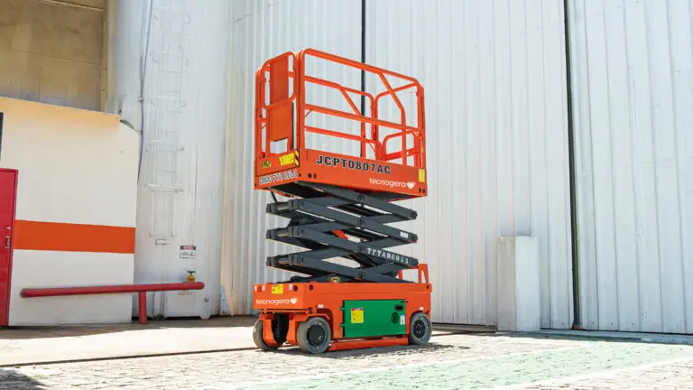 Uma plataforma elevatória tipo tesoura, na cor laranja vibrante, está totalmente elevada em uma área externa de concreto, em frente a um grande galpão industrial branco. O equipamento possui apoios e grades pretos e representa uma das principais soluções da Tecnogera para trabalhos em altura.