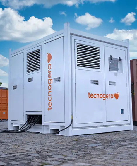 Um grande gerador industrial branco com o nome Tecnogera está posicionado sobre um piso de paralelepípedos, com diversos cabos pretos saindo da sua base. Ao fundo, é possível ver um container laranja e equipamentos para trabalhos em altura, tudo sob um céu parcialmente nublado.