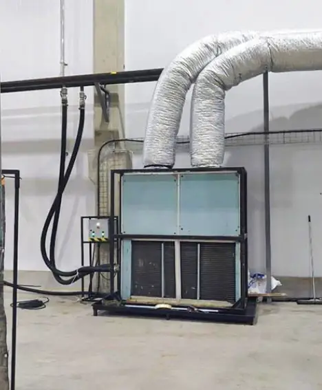 Um grande equipamento de ar-condicionado industrial com dois dutos prateados isolados se eleva para o alto. Perto dele, estão painéis de controle e tubulações pretas, enquanto produtos de limpeza e plataformas para trabalhos em altura da Tecnogera apoiam-se na parede branca ao fundo.