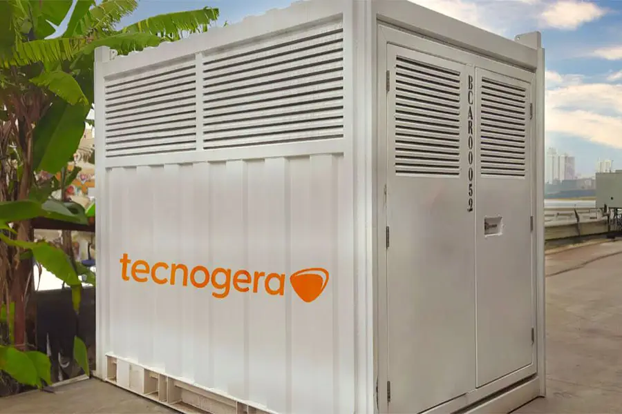 Um grande contêiner industrial branco, com aberturas de ventilação e uma porta, está posicionado ao ar livre próximo a plantas verdes. O logotipo da Tecnogera, em laranja, aparece destacado na lateral. Ao fundo, sob um céu nublado, é possível ver o horizonte da cidade e plataformas para trabalhos em altura.
