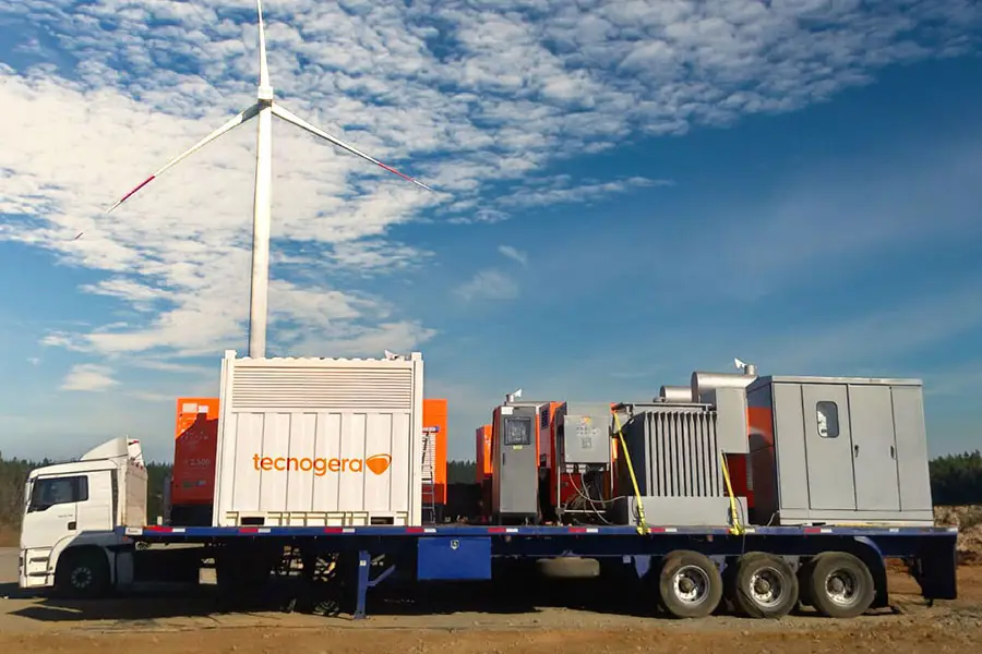 Um caminhão branco com carreta azul transporta equipamentos industriais da Tecnogera, especialista em plataformas para trabalhos em altura. Ao fundo, um aerogerador se destaca no céu parcialmente nublado, sobre um solo de terra e vegetação ao longe.