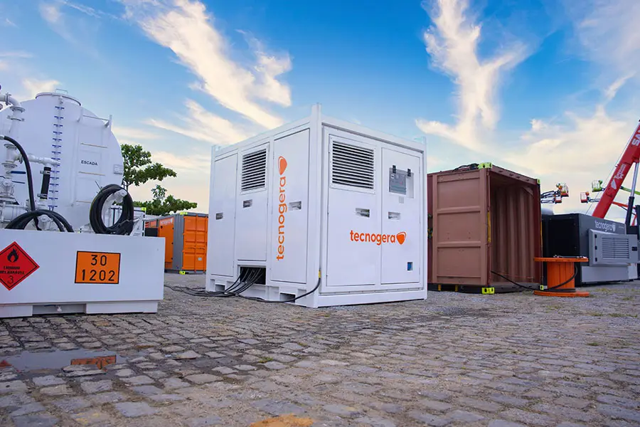 Um banco de carga branco da Tecnogera está posicionado sobre um piso de paralelepípedos ao ar livre, cercado por equipamentos industriais, contêineres, plataformas para trabalho em altura e outras máquinas. O cenário é um pátio ou área de armazenamento sob um céu azul com algumas nuvens finas.