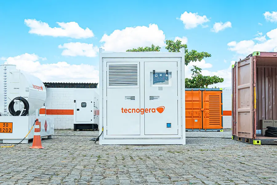 Um gerador industrial branco da Tecnogera está posicionado em um pátio de paralelepípedos, sob um céu azul com algumas nuvens. Perto dele, há um carretel, um cone de sinalização e um container laranja-um cenário perfeito para o uso de plataformas para trabalhos em altura. Ao fundo, é possível ver uma árvore e um muro.