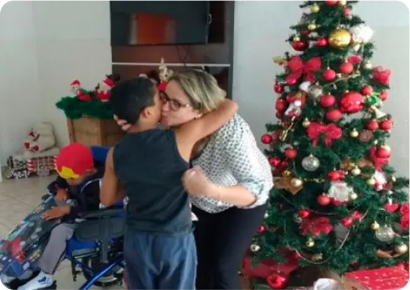 Uma mulher abraça um menino em frente a uma árvore de Natal decorada com enfeites vermelhos e dourados. Perto deles, outra criança em uma cadeira de rodas, usando um gorro vermelho, faz parte da cena-uma imagem que transmite acolhimento e inclusão com o jeito Tecnogera de ser. Os presentes ao redor completam o clima alegre e festivo.