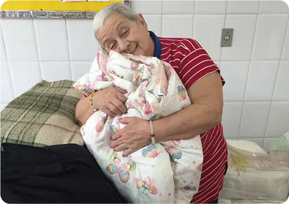 Uma mulher mais velha, de cabelos grisalhos curtos e usando uma camisa listrada vermelha, sorri de olhos fechados enquanto abraça um cobertor dobrado e colorido com estampas de borboleta. Ela está ao lado de uma cama coberta por mantas estampadas, transmitindo aconchego junto à Tecnogera.