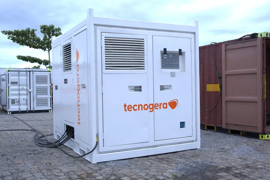 Um grande gerador industrial branco, com o nome Tecnogera, está instalado ao ar livre sobre um piso pavimentado, com cabos conectados à sua lateral. Ao redor, é possível ver contêineres, árvores e plataformas para trabalhos em altura sob um céu nublado.