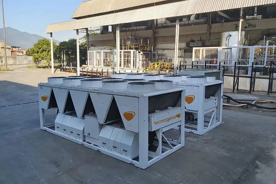 Dois grandes aparelhos industriais de ar-condicionado branco, com ventiladores visíveis no topo e o logotipo da Tecnogera, estão posicionados ao ar livre sobre uma superfície de concreto. Próximo a eles, plataformas para trabalho em altura auxiliam na manutenção de tubulações, estruturas metálicas e do prédio industrial ao redor.