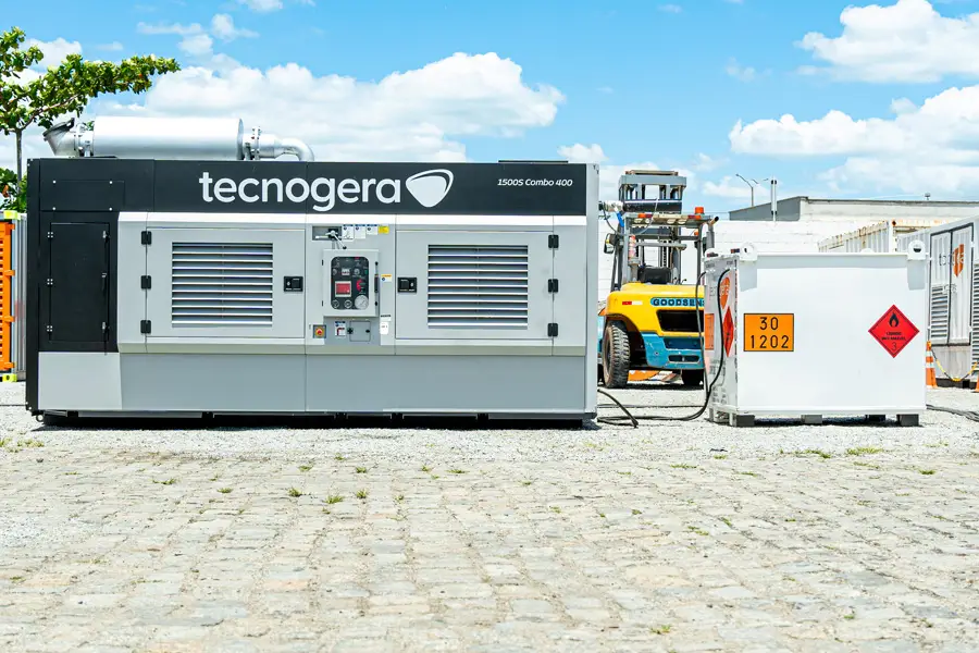 Um grande gerador cinza e preto da Tecnogera, identificado como "1500k Cambio 4009", está posicionado sobre um piso pavimentado ao lado de um tanque de combustível branco com etiquetas de segurança. Ao fundo, sob o céu azul, é possível ver uma empilhadeira amarela, plataformas para trabalho em altura e outros equipamentos.