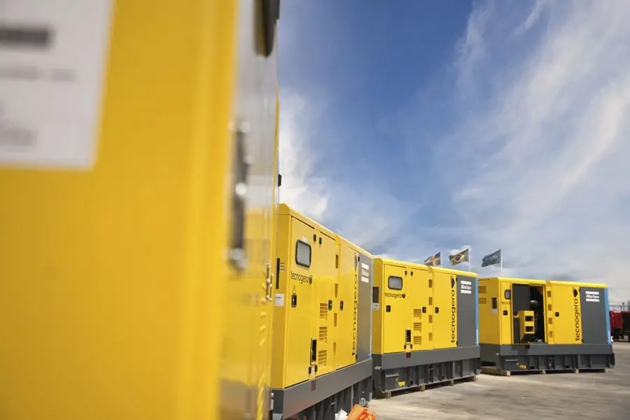 Grandes geradores industriais amarelos da Tecnogera estão alinhados ao ar livre, sob um céu azul com algumas nuvens, ao lado de plataformas para trabalhos em altura. A foto, tirada em um ângulo lateral, destaca as laterais e portas dos equipamentos, enquanto bandeiras aparecem ao fundo.
