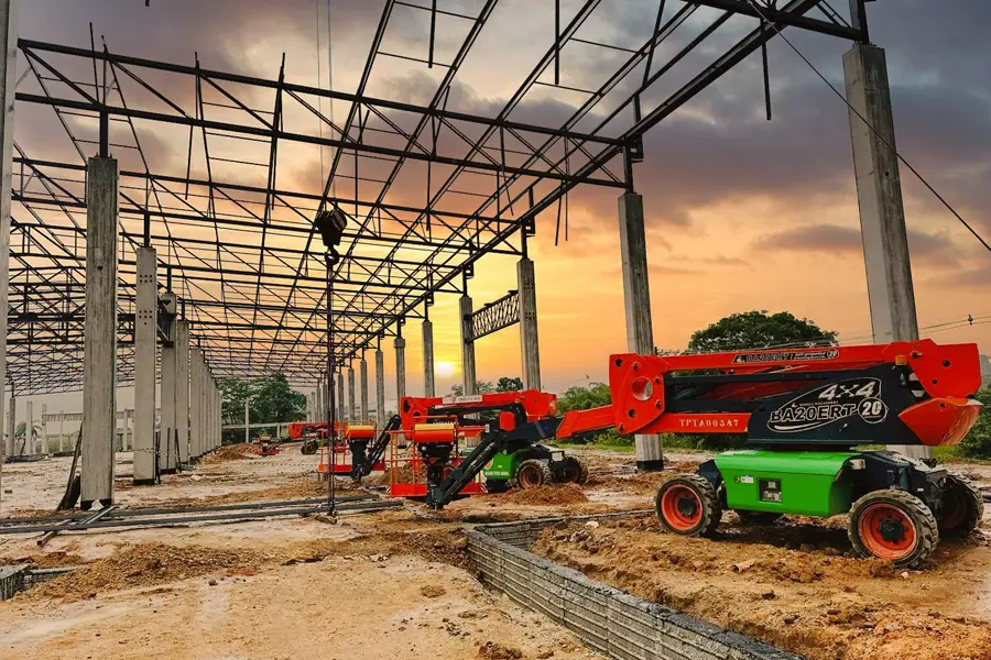 Obra ao entardecer, destacando a estrutura de aço e a base de concreto de um grande edifício. Três plataformas para trabalho em altura da Tecnogera (duas vermelhas e uma verde) estão posicionadas sobre o solo úmido, sob um céu colorido em tons de laranja e roxo.