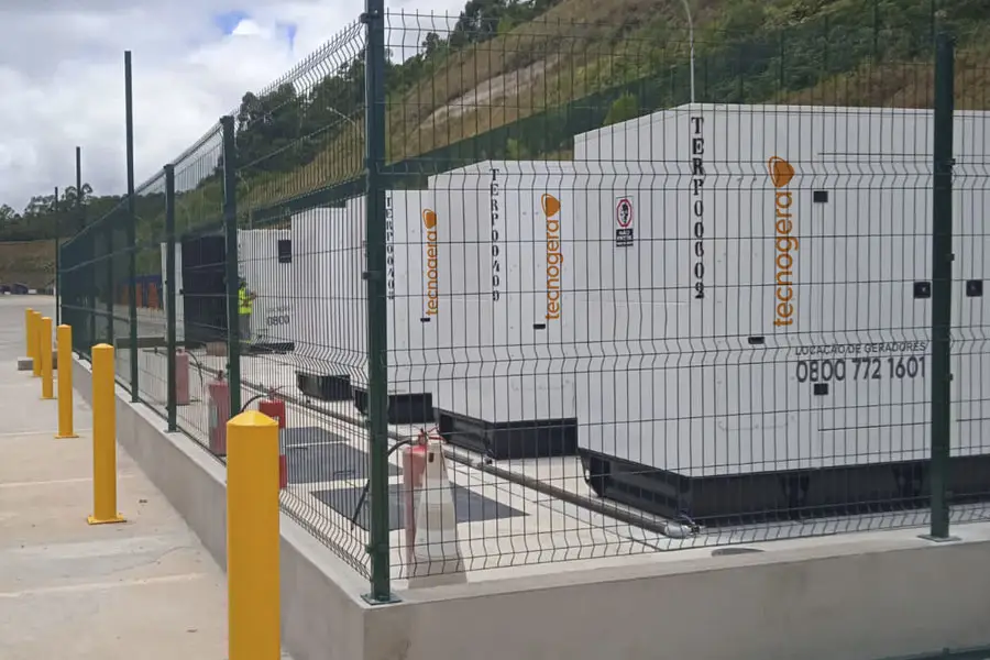 Vários grandes geradores industriais brancos, identificados com a marca Tecnogera, estão protegidos por uma alta cerca de metal com arame farpado. No espaço de concreto ao redor, há balizadores amarelos para segurança e plataformas para trabalhos em altura. Ao fundo, é possível ver colinas cobertas de grama.