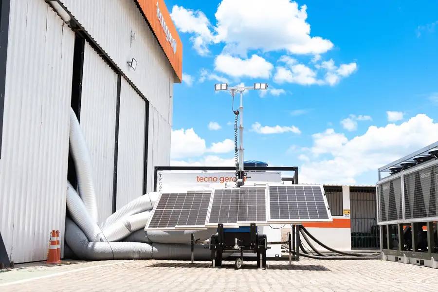 Uma unidade móvel de painéis solares com grandes placas está instalada do lado de fora de um prédio industrial sob um céu azul. No local, há dutos flexíveis e equipamentos com a marca "tecno gerdau", além de dois cones laranja e plataformas para trabalho em altura da Tecnogera.