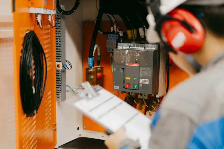 Na imagem, um profissional equipado com EPI, como capacete e protetor auricular vermelho, inspeciona um quadro elétrico aberto próximo às plataformas para trabalho em altura. Ele segura uma prancheta e observa os disjuntores, fios e componentes dentro do painel laranja. Esta cena mostra o cuidado com a segurança que a Tecnogera adota em suas operações.