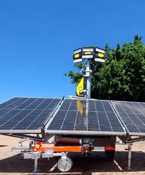 Um trailer solar móvel da Tecnogera, equipado com grandes painéis solares e refletores, funciona como uma torre de iluminação. Ele está estacionado sobre o asfalto em um dia ensolarado, com árvores verdes ao fundo. O trailer conta ainda com rodas e suportes metálicos para garantir estabilidade e praticidade no uso.
