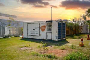 Um contêiner branco com a marca Tecnogera está posicionado sobre a grama, sob um céu de pôr do sol, ilustrando os desafios da organização de eventos: como garantir estrutura adequada e fornecimento contínuo de energia em um ambiente calmo e industrial. Pequenas árvores e extintores de incêndio completam o cenário ao redor.