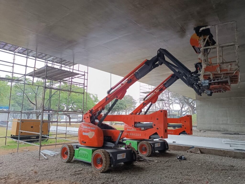Dois elevadores articulados vermelhos da Tecnogera estão posicionados sob uma ponte de concreto, auxiliando na manutenção predial com segurança e eficiência. Um dos equipamentos está estendido para cima, permitindo que um colaborador alcance a parte inferior da estrutura, enquanto andaimes e árvores completam o cenário sobre o solo de cascalho.