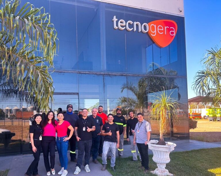 Um grupo diverso de doze pessoas sorri em frente a um prédio moderno de vidro com a placa da Tecnogera, mostrando que a Tecnogera é muito mais do que uma simples locadora de geradores. Palavras decoram o ambiente ensolarado, onde palmeiras e um vaso branco completam o cenário.