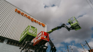 Duas plataformas elevatórias verdes alcançam um galpão de metal ondulado, que exibe o logotipo laranja da Tecnogera na fachada. O céu está nublado, fios elétricos aparecem ao fundo e uma das plataformas sustenta um cesto vermelho. Tecnogera: muito mais do que locação de geradores.