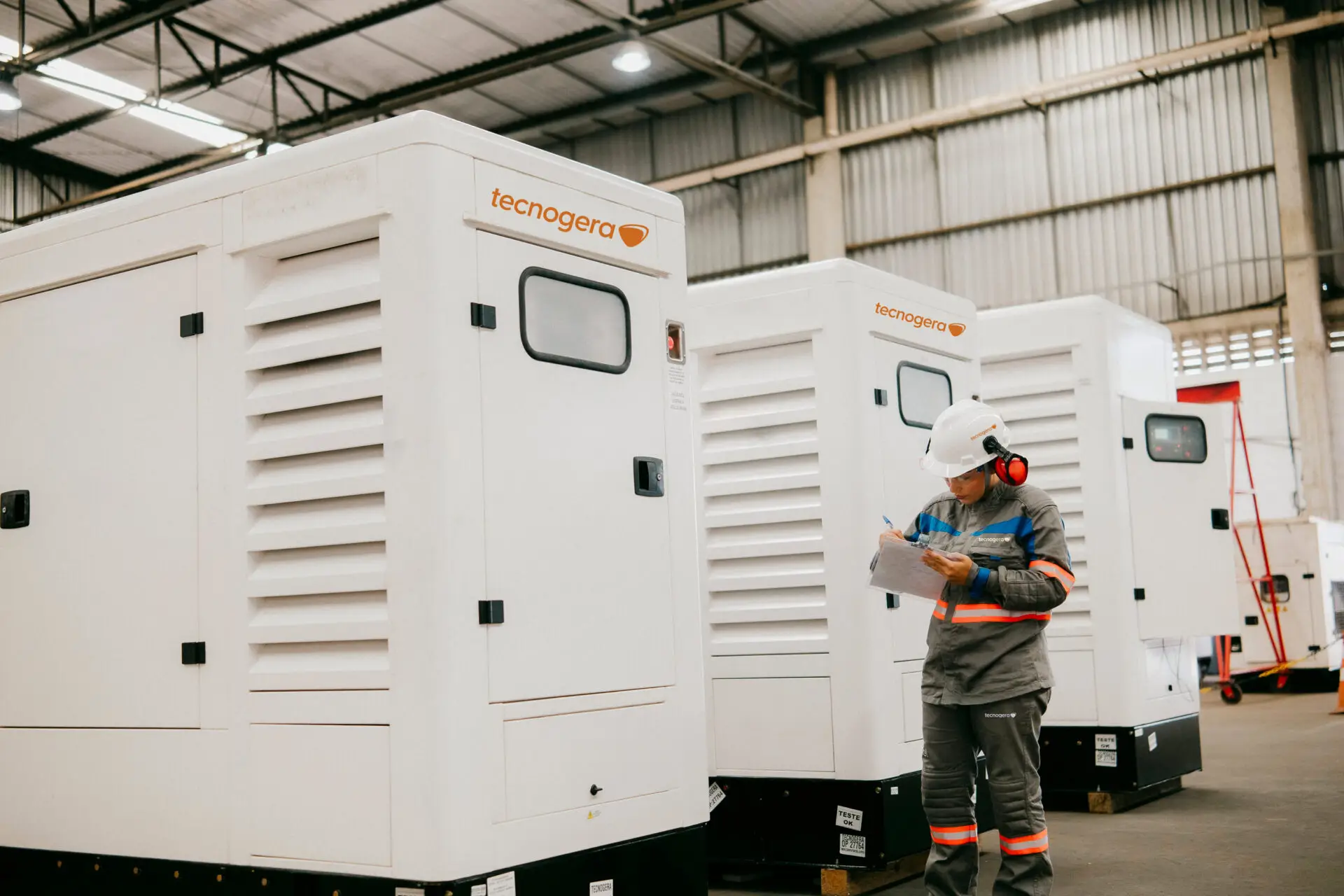 Um colaborador equipado com itens de segurança, como capacete, luvas e proteção auricular, confere documentos ao lado dos grandes geradores da Tecnogera, garantindo energia eficiente para eventos corporativos em um galpão amplo e iluminado.