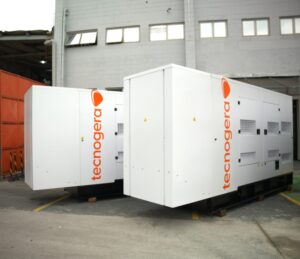 Dois geradores grandes da Tecnogera, estão posicionados sobre paletes ao ar livre, próximos a um prédio cinza. Perfeitos para garantir operações eficientes.