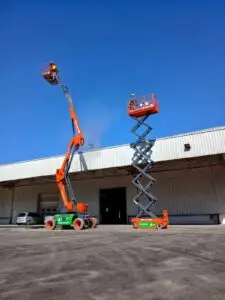 Na imagem, dois profissionais da Tecnogera usam EPIs e operam plataformas elevatórias laranja próximas a um prédio industrial.