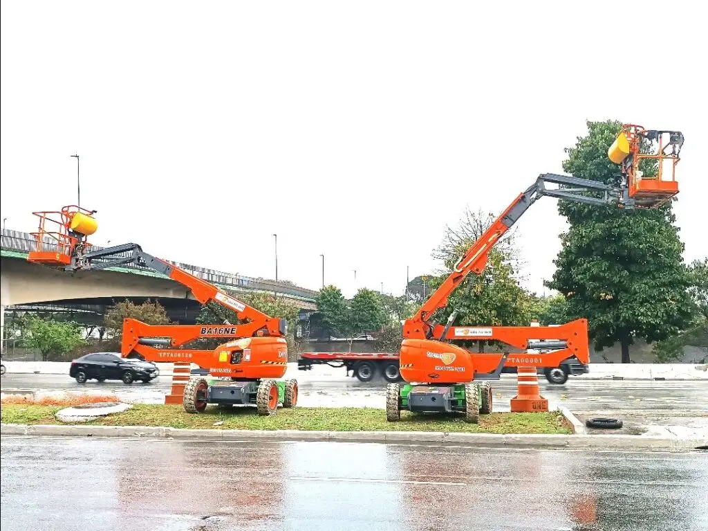 Na imagem, plataformas elevatórias da Tecnogera operam na chuva, sob céu nublado próximo a uma ponte.
